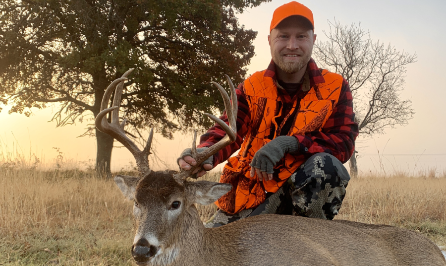 A hunter wearing an orange safety vest and cap smiles proudly while kneeling beside a harvested buck with impressive antlers.