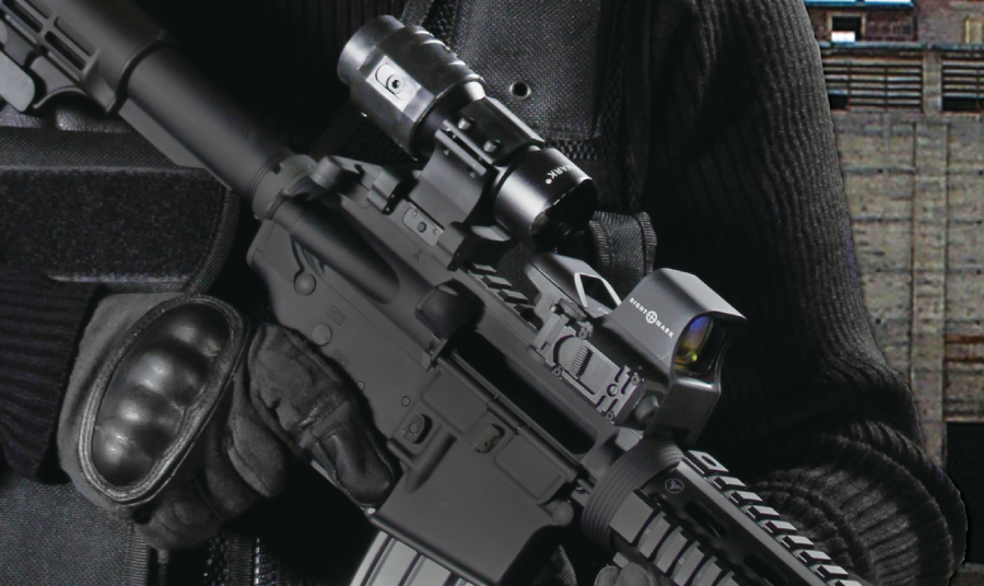 The image shows a close-up of a tactical rifle equipped with multiple optics, including a red dot sight and a magnifier.